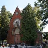 Ceglana kaplica stojąca przy alejce na cmentarzu, częściowo zasłonięta przez drzewa.