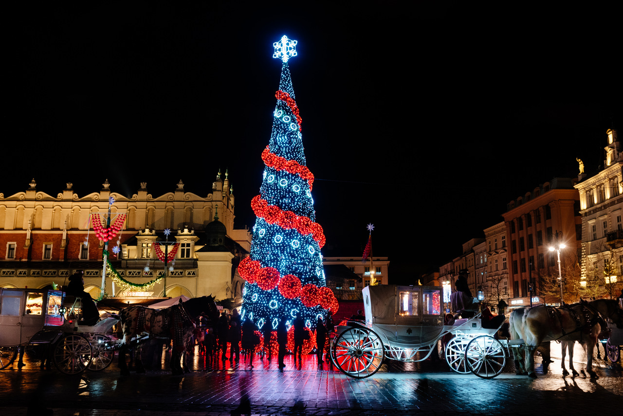 In der Mitte steht ein beleuchteter Weihnachtsbaum auf dem Hauptplatz in Krakau. Eine Kutsche mit Pferden fährt vorbei. Im Hintergrund die Tuchhallen und Mietshäuser bei Nacht.