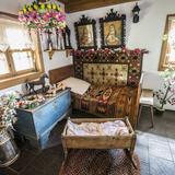 Wnętrze drewnianej chaty z malowana skrzynią, kołyska, łóżkiem z kolorową narzutą i poducha, makatą i obrazami świętych na ścianach, bibułowymi pająkami i kwiatami, koronkowymi firankami w oknie.
