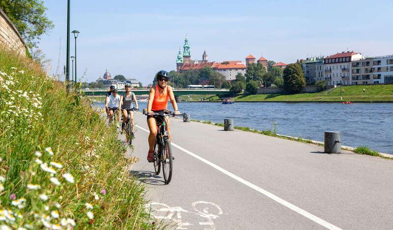 Rowerzyści jada po ścieżce rowerowej nad rzeką w tle widać Wawel
