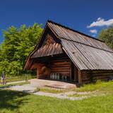 Drewniana Bacówka Wojciecha Komperdy w Czorsztynie. Obok niej trzech turystów przechodzących ścieżką. Wokół bacówki sporo zieleni, drzew i niebieskie niebo nad nimi.