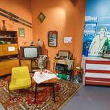 Aranżacja pokoju z czasów PRL jako ekspozycja w Muzeum Niepodległości w Myślenicach. Znajduje się w nim komoda, telewizor na półce, fotel i stolik, a także drobniejsze rzeczy. Po prawej stronie natomiast widać wagę sklepową i plakat.