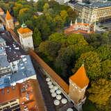 Kawałek murów obronnych z wieżami w Krakowie widzianych z lotu ptaka. Po lewej widać zabudowania kamienic Starego Miasta, po prawej od murów znajdują się drzewa oraz okrąg z ceglanych murów z siedmioma wieżyczkami i z kwadratową długą bramą wjazdową. Dalej za nimi stare, wysokie kamienice.