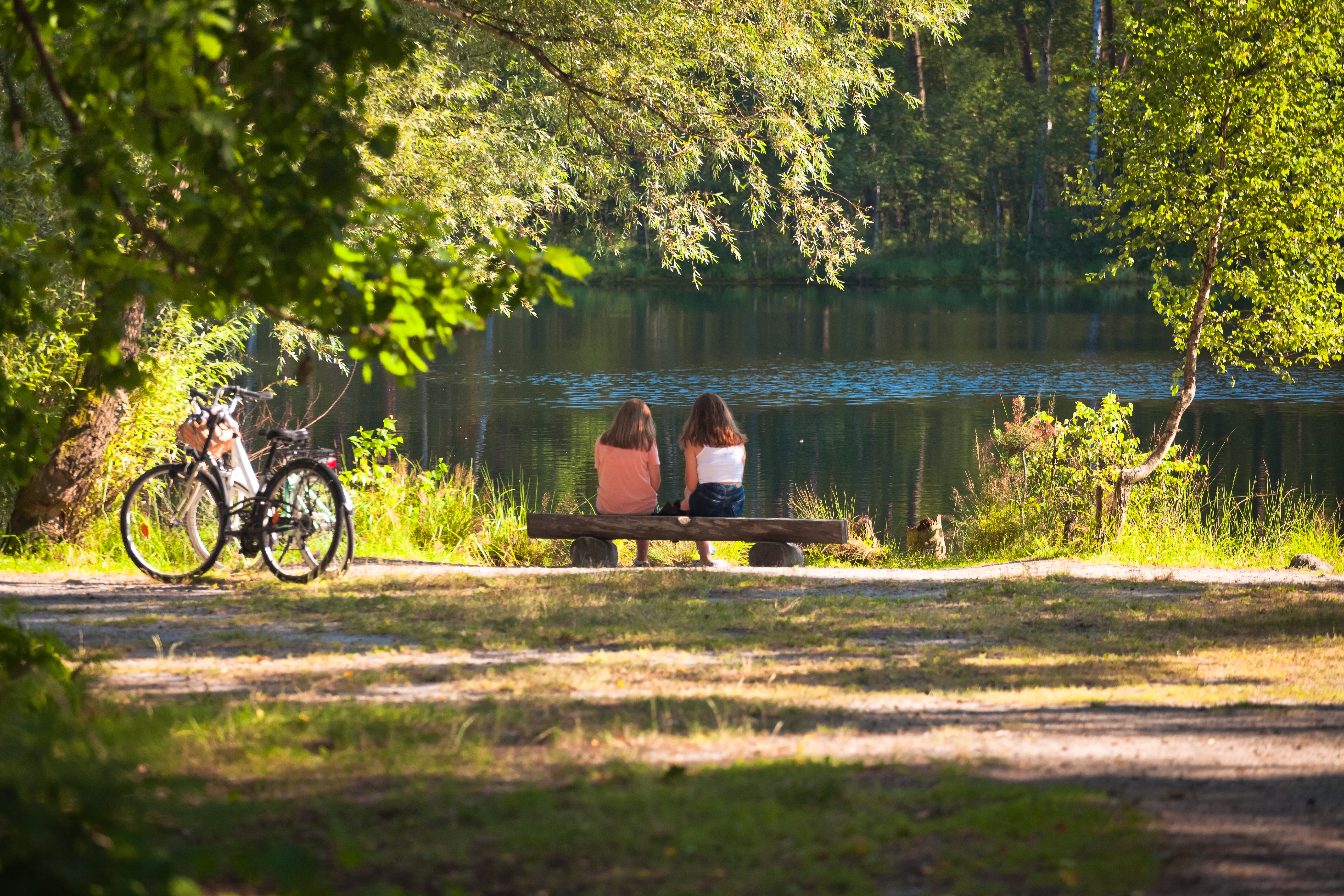 Dwie dziewczyny siedzą na ławce nad stawem w lesie, obok stoją zaparkowane rowery